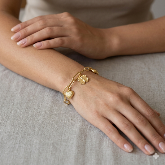 Gold bracelet with heart and clover charms on a wrist against a neutral background