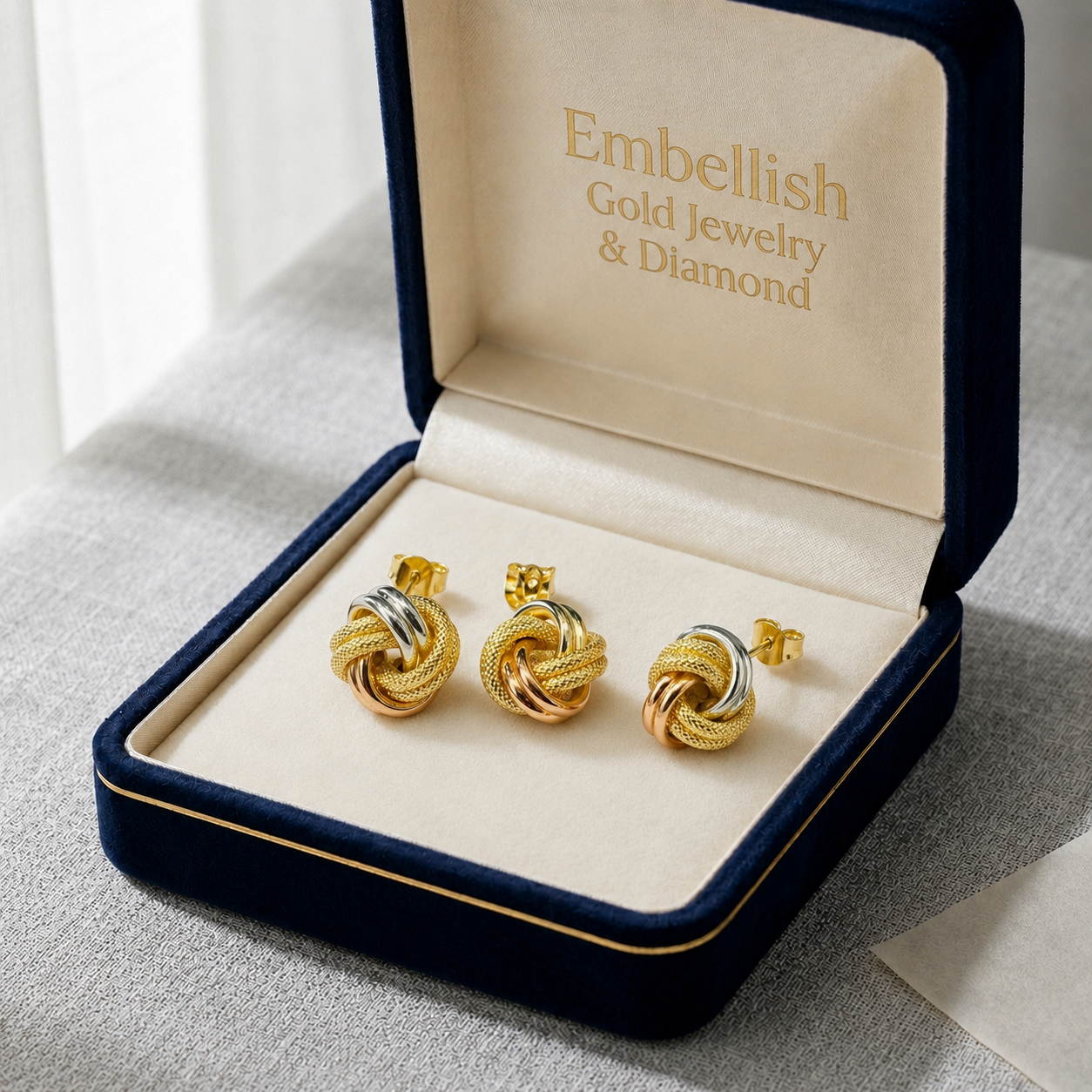 Gold knot earrings in a 'Embellish Gold Jewelry & Diamond' box on a gray surface.