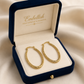 Gold hoop earrings in a 'Embellish' jewelry box on a beige fabric background