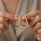 Gold triangle-shaped earrings held between two hands against a neutral background