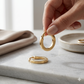 Gold hoop earrings being held by a hand on a marble surface with a neutral background